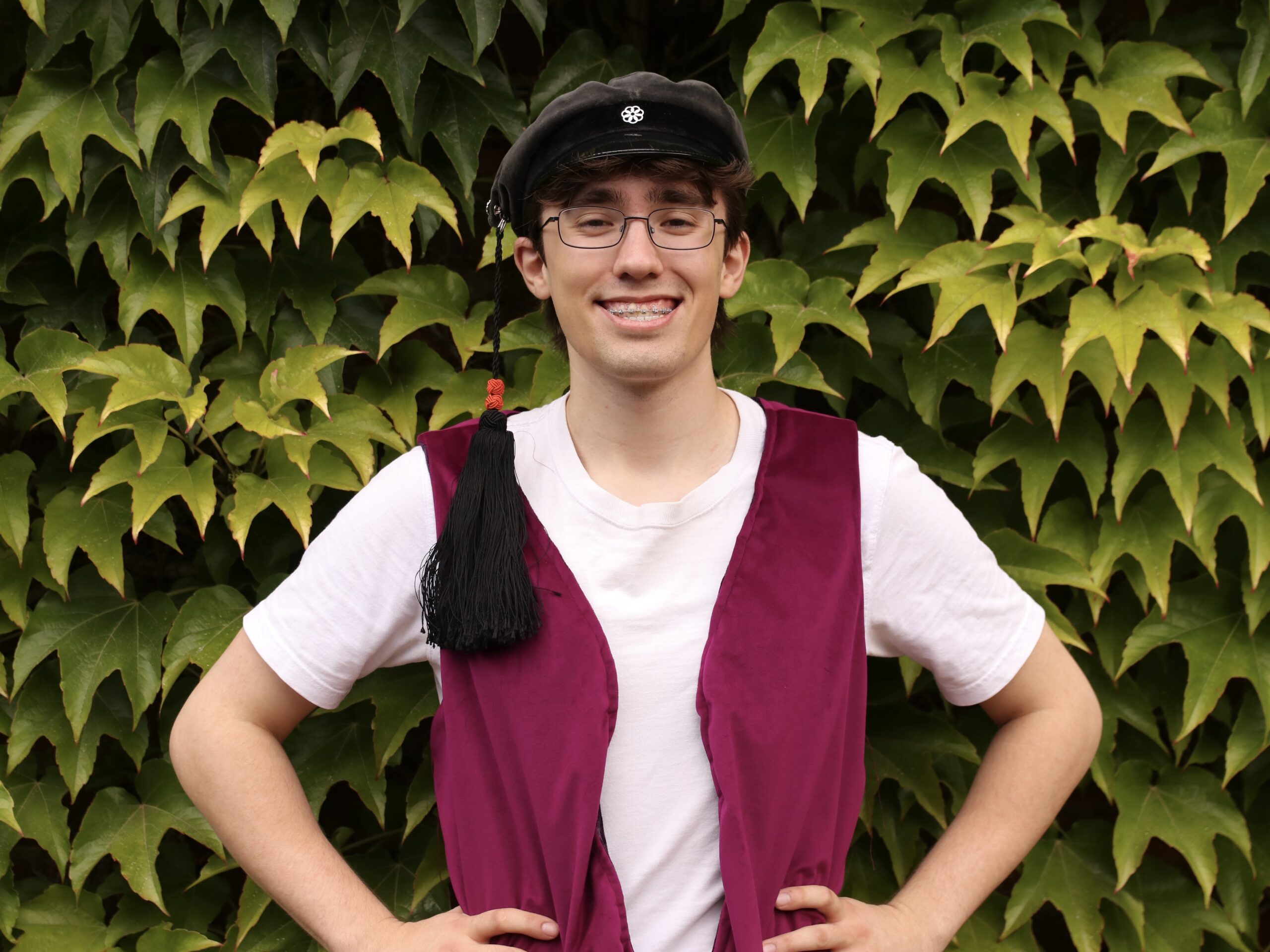 David har en vit T-shirt och en cerise väst och står framför en vägg klädd med murgröna. David har glasögon, kort brunt hår under en svart schmeck och ler brett.